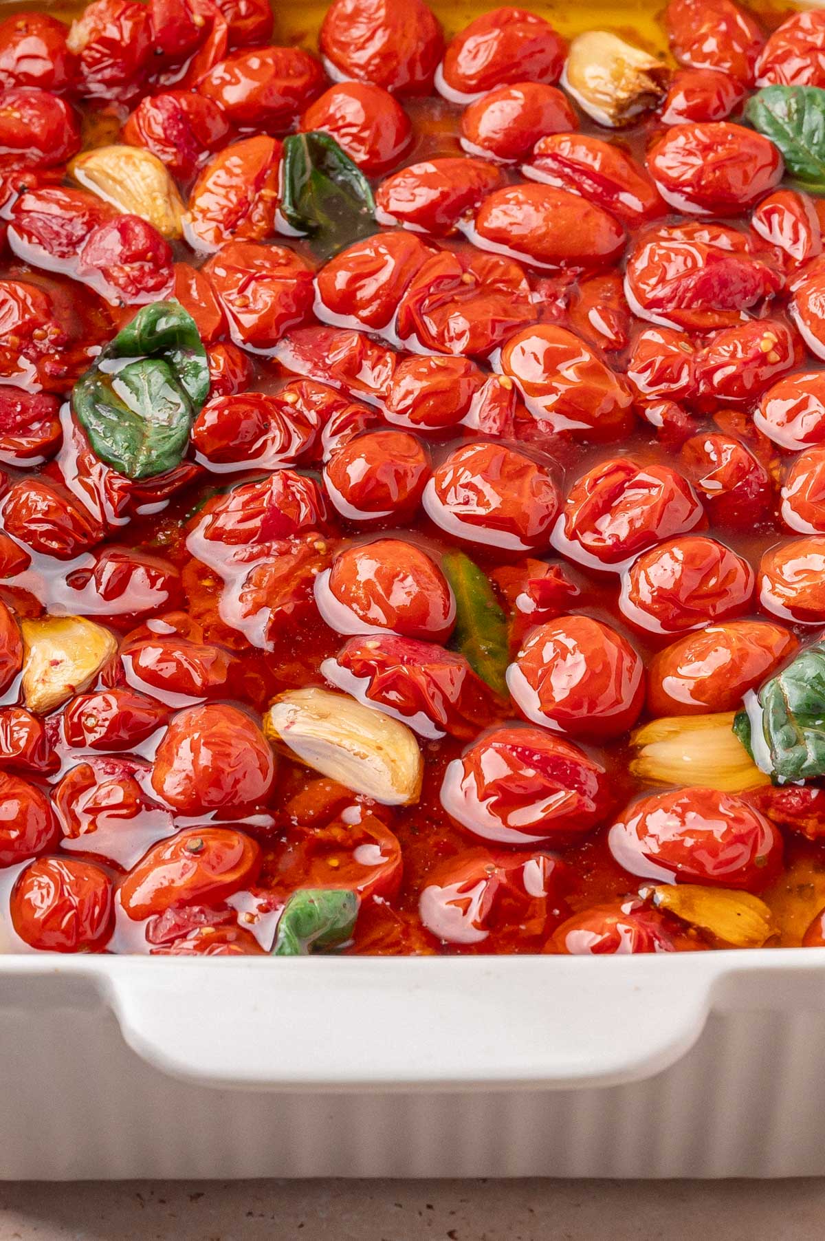 Glossy, oil-soaked roasted cherry tomatoes with garlic and basil in a white dish.
