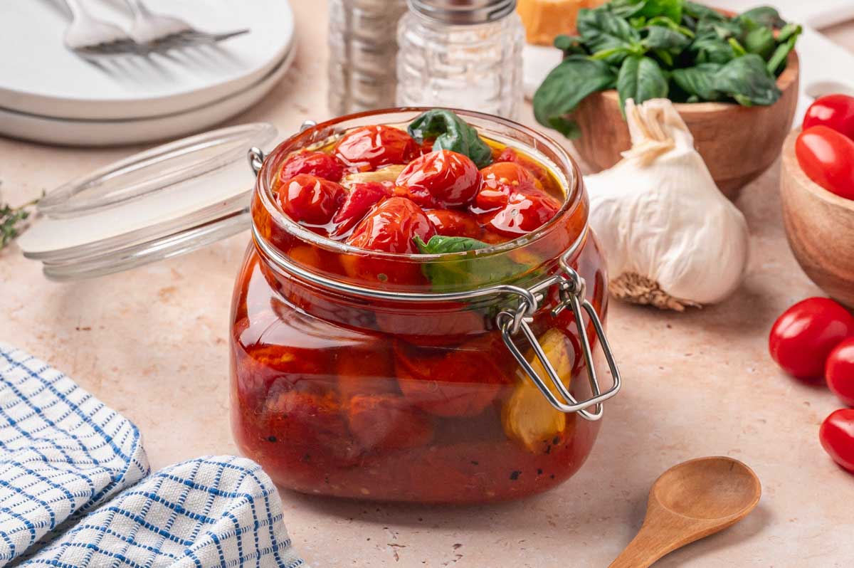 Cherry tomato confit stored in a glass jar with garlic and basil, surrounded by ingredients.