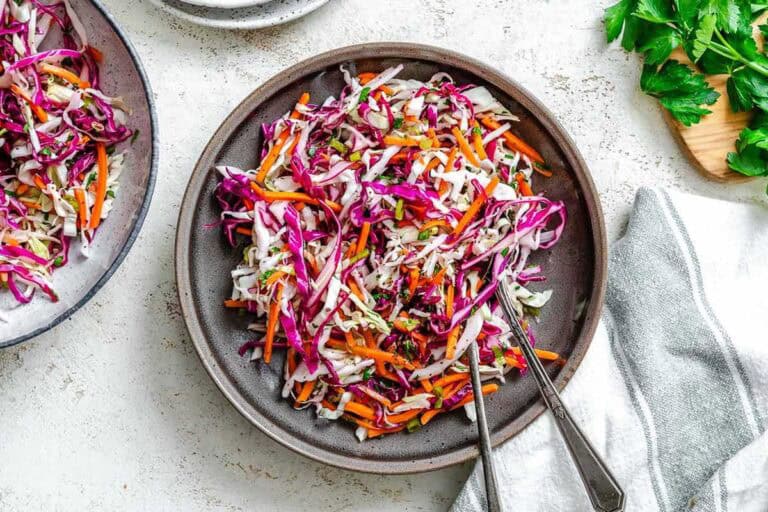 A colorful cabbage slaw with shredded red and green cabbage, carrots, and fresh herbs served in a ceramic brown bowl with silver serving utensils.