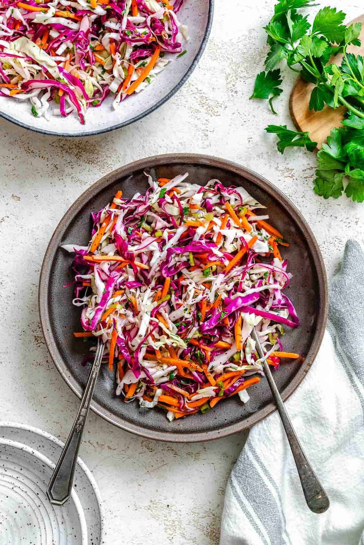 A finished bowl of vibrant vinegar slaw surrounded by fresh parsley, an empty plate, and a folded towel on a white textured surface.
