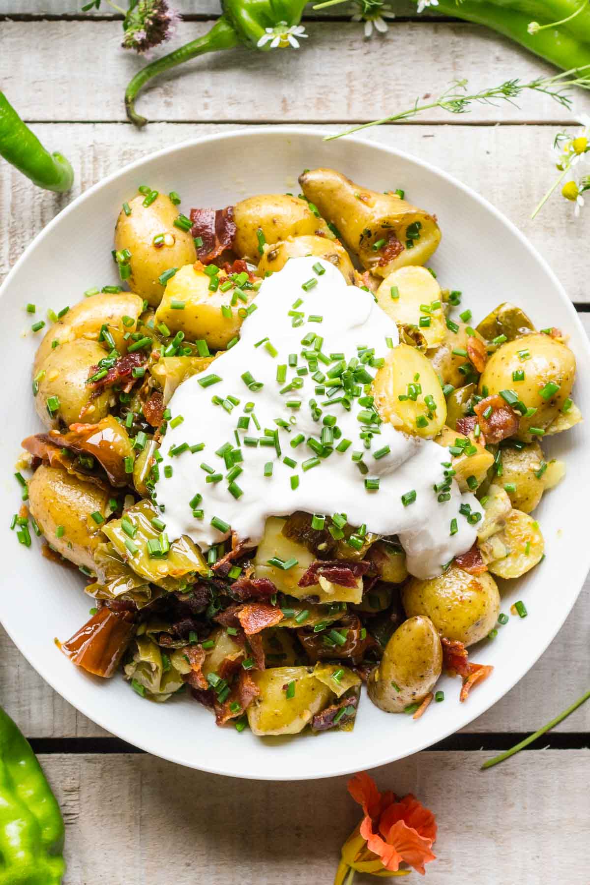 Overhead view of a white bowl filled with warm potato salad, topped with crispy bacon, sautéed peppers, and a generous dollop of sour cream, sprinkled with chopped chives.