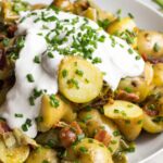 Angled close-up of potato salad with creamy sour cream and fresh chives on top, highlighting the sliced fingerling potatoes and crispy bacon bits.