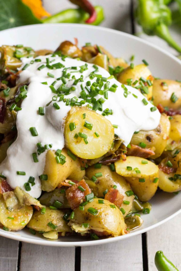 Angled close-up of potato salad with creamy sour cream and fresh chives on top, highlighting the sliced fingerling potatoes and crispy bacon bits.
