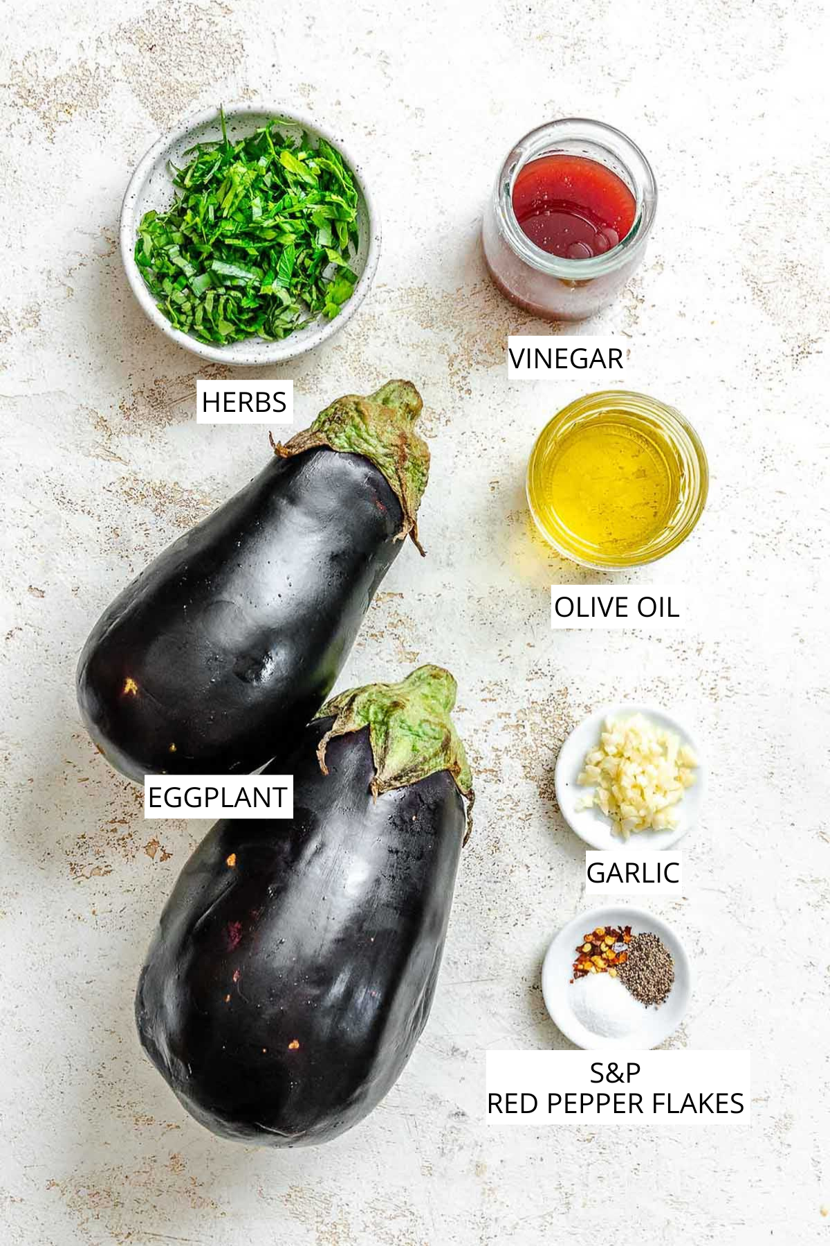 Overhead view of ingredients for grilled eggplant, including two whole eggplants, chopped fresh herbs, red vinegar, olive oil, minced garlic, and a small dish of salt, pepper, and red pepper flakes, each clearly labeled with text.