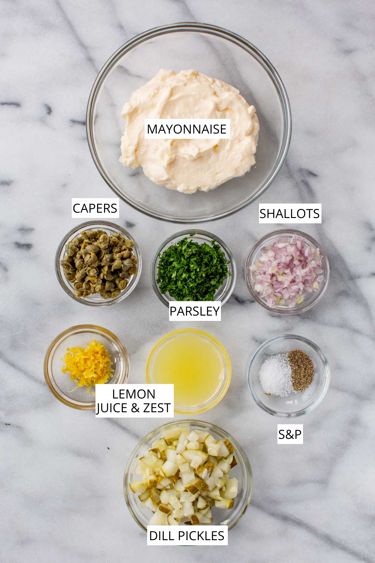 Labeled ingredients for tartar sauce arranged on a marble surface, including bowls of mayonnaise, chopped capers, parsley, shallots, dill pickles, lemon juice and zest, and salt and pepper.