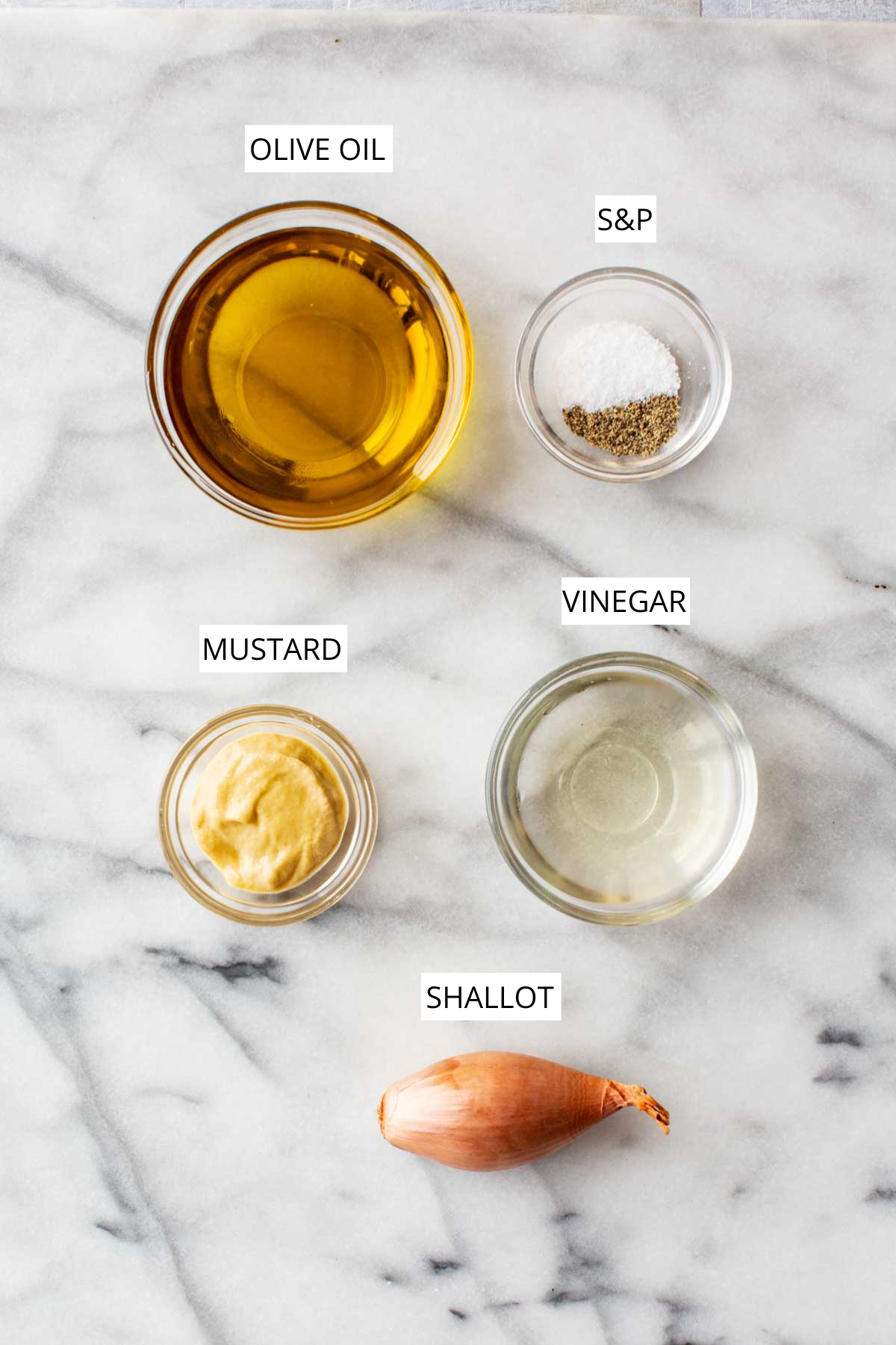 Flat lay of vinaigrette ingredients labeled on a marble background: olive oil, salt and pepper, white wine vinegar, Dijon mustard, and shallot.
