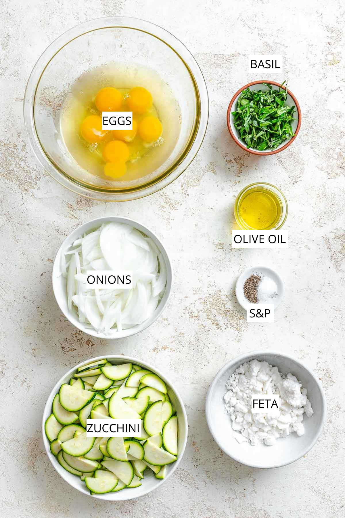 Overhead shot of ingredients including eggs, onions, zucchini, feta, basil, olive oil, and salt and pepper, arranged neatly on a white surface.