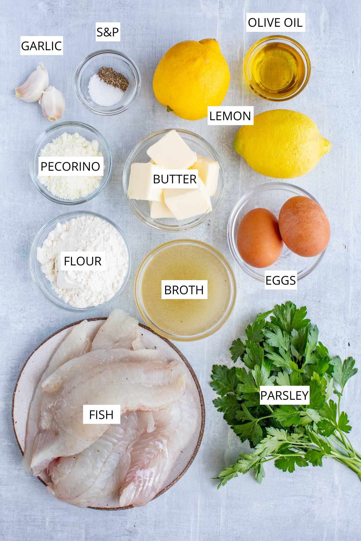Overhead shot of labeled ingredients for flounder francaise including fresh flounder filets, eggs, lemon, parsley, broth, butter, flour, pecorino, garlic, olive oil, and salt and pepper.