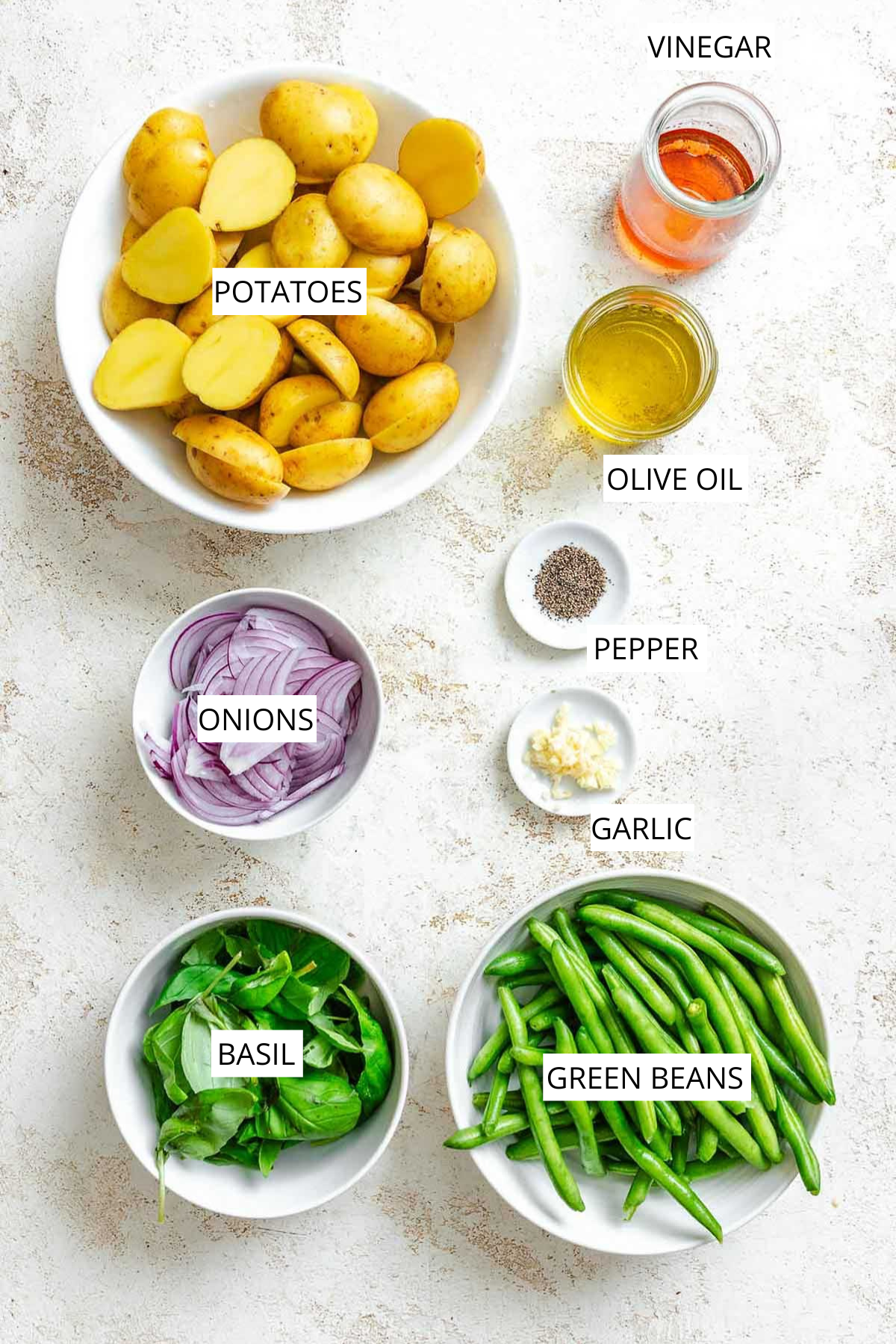 Ingredients for the salad displayed in separate bowls: halved yellow potatoes, red wine vinegar, olive oil, garlic, sliced red onions, green beans, and fresh basil.