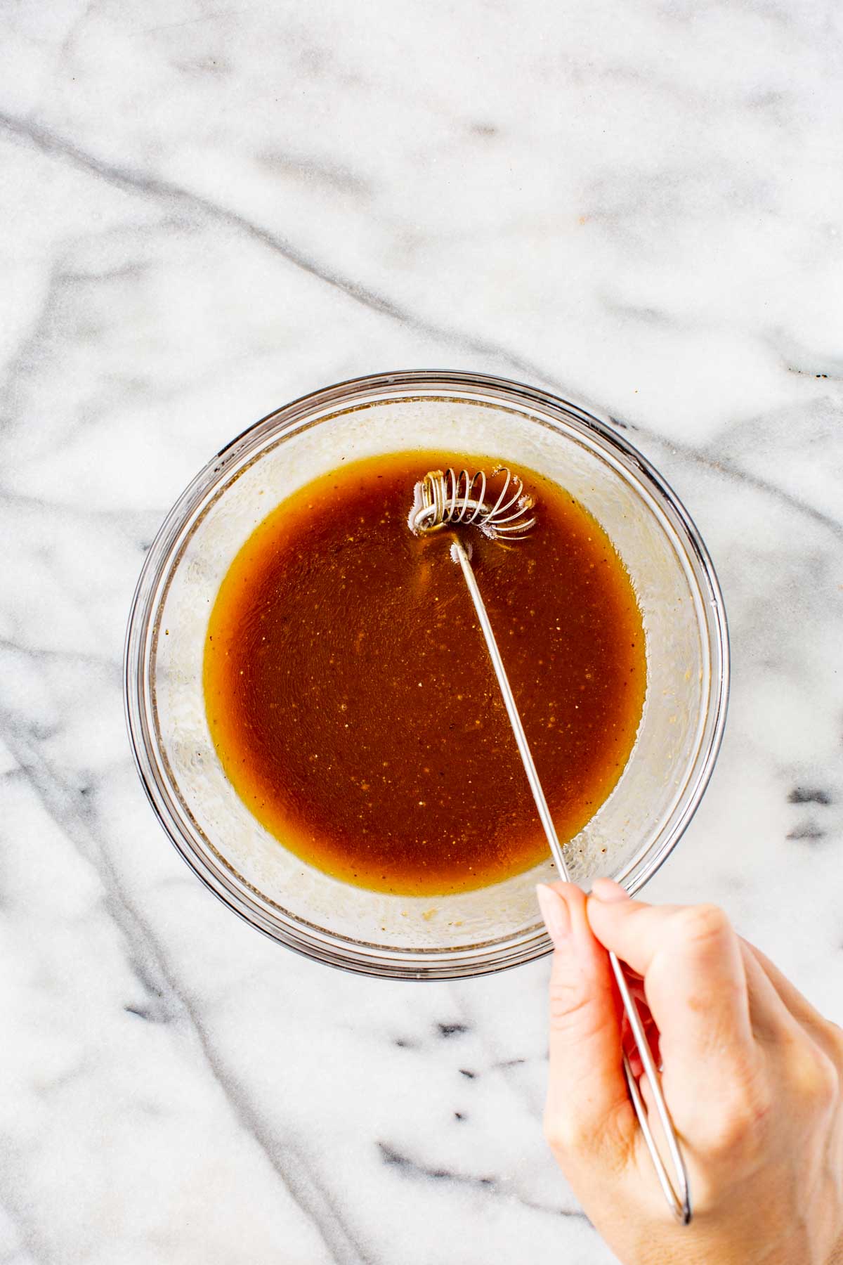 Whisking balsamic vinaigrette until fully emulsified in a clear glass bowl on a marble surface.