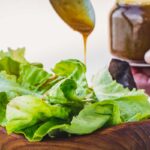 Drizzling balsamic vinaigrette over fresh lettuce in a wooden bowl with a hand holding the jar in the background.