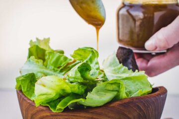 Drizzling balsamic vinaigrette over fresh lettuce in a wooden bowl with a hand holding the jar in the background.