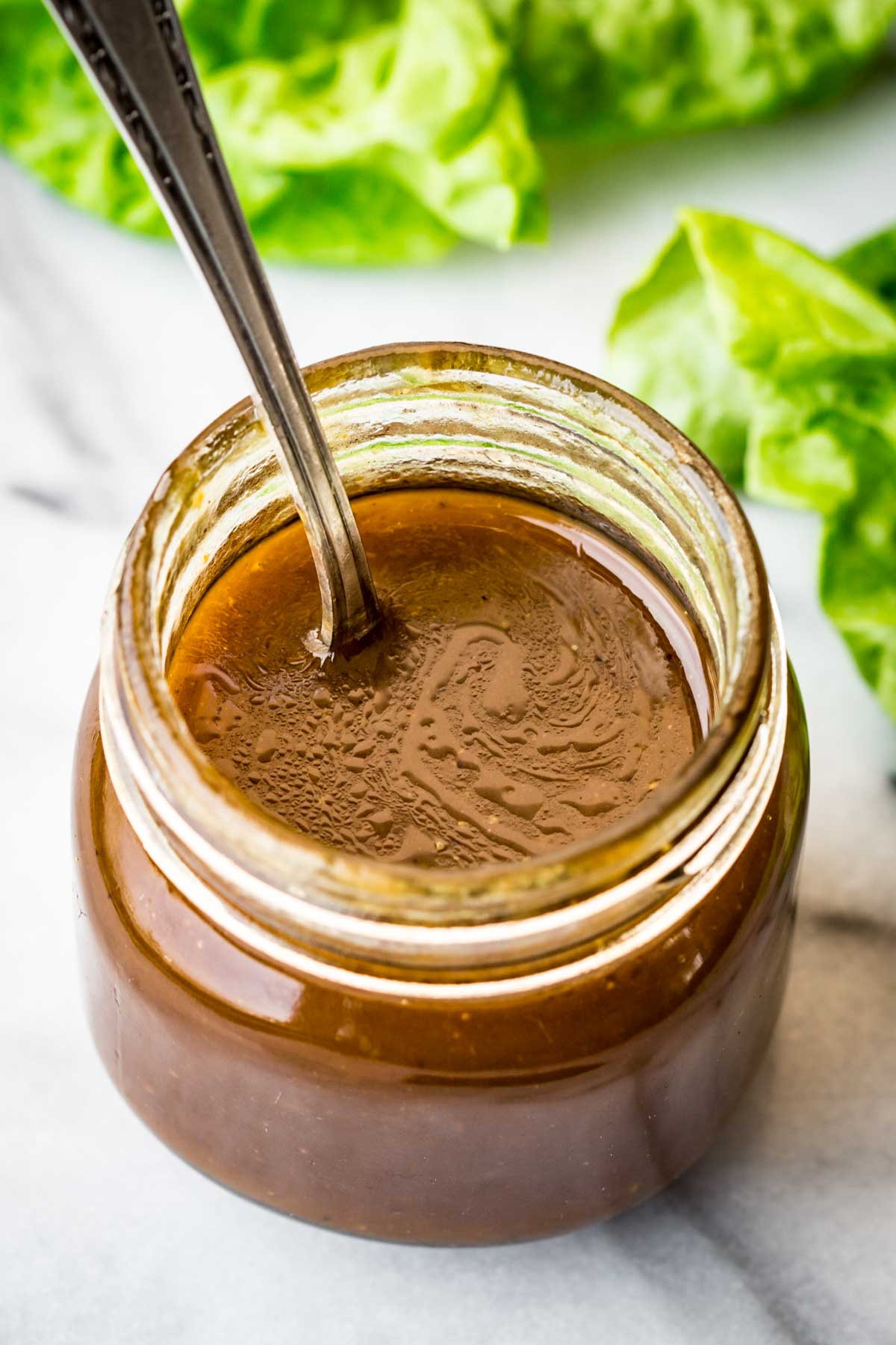 Overhead shot of an open jar of balsamic vinaigrette with a spoon inside, surrounded by fresh lettuce leaves.