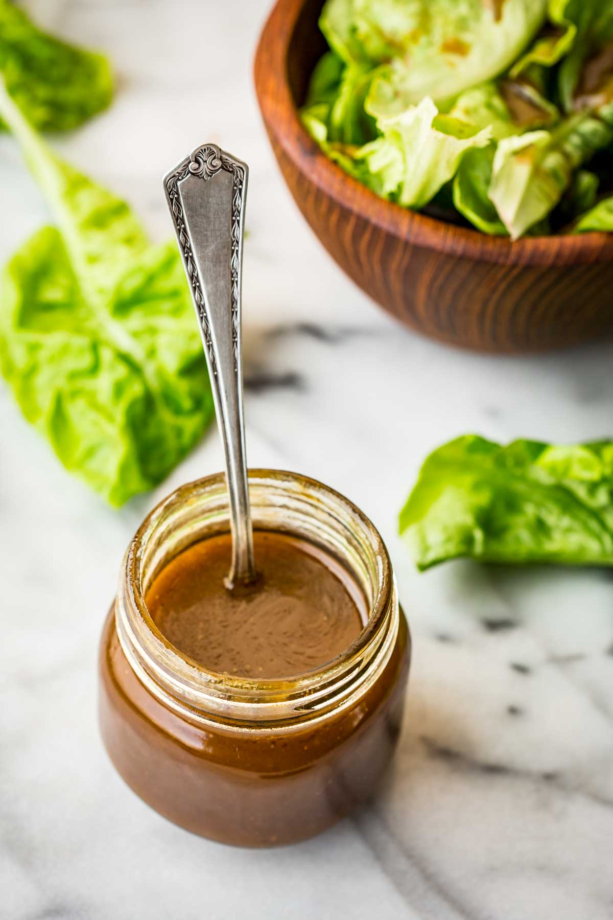Glass jar of balsamic vinaigrette with spoon standing upright, set next to a salad bowl and scattered greens.