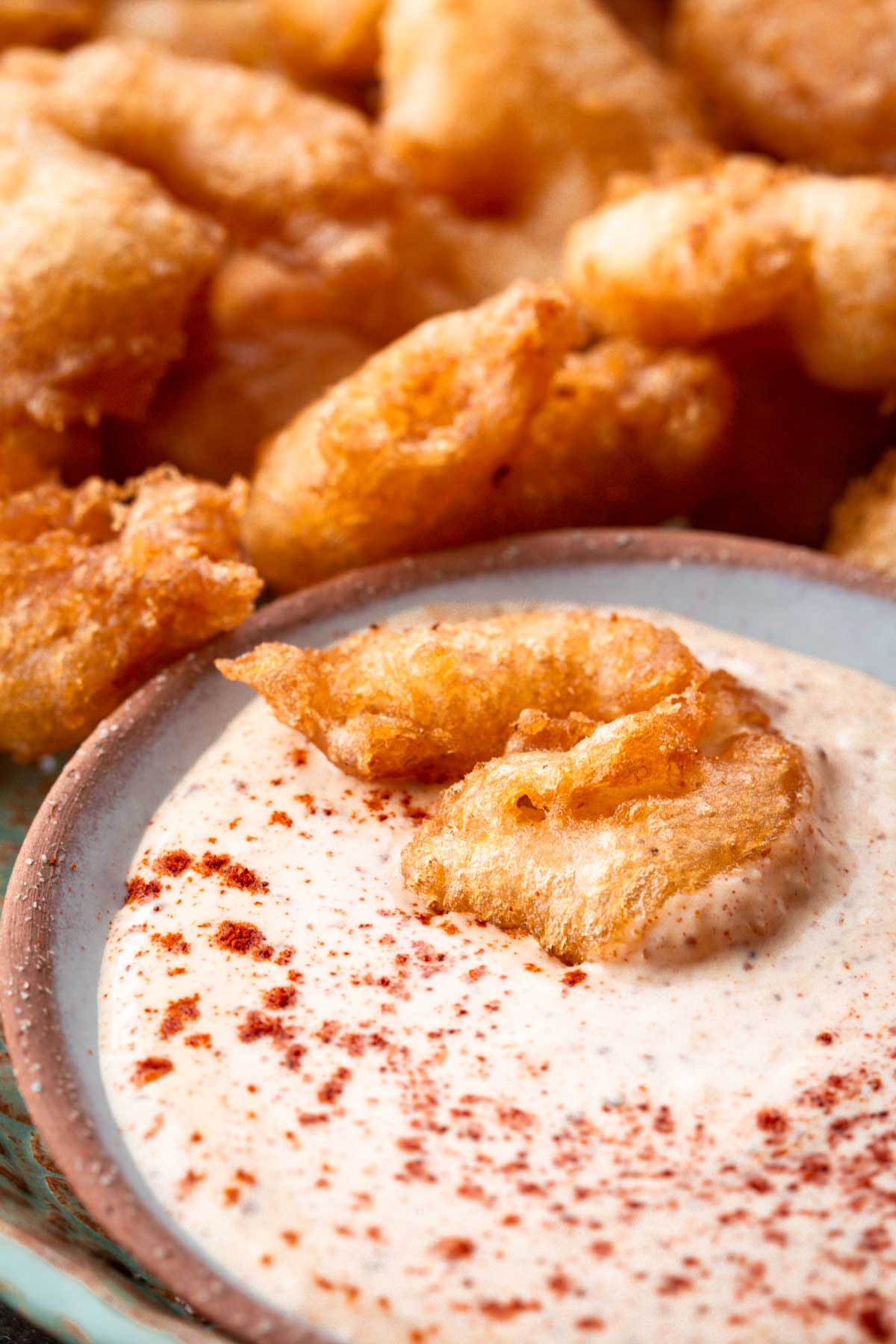 Extreme close-up of a crispy, golden beer battered shrimp partially dipped in creamy, paprika-dusted dipping sauce with more shrimp in the background.