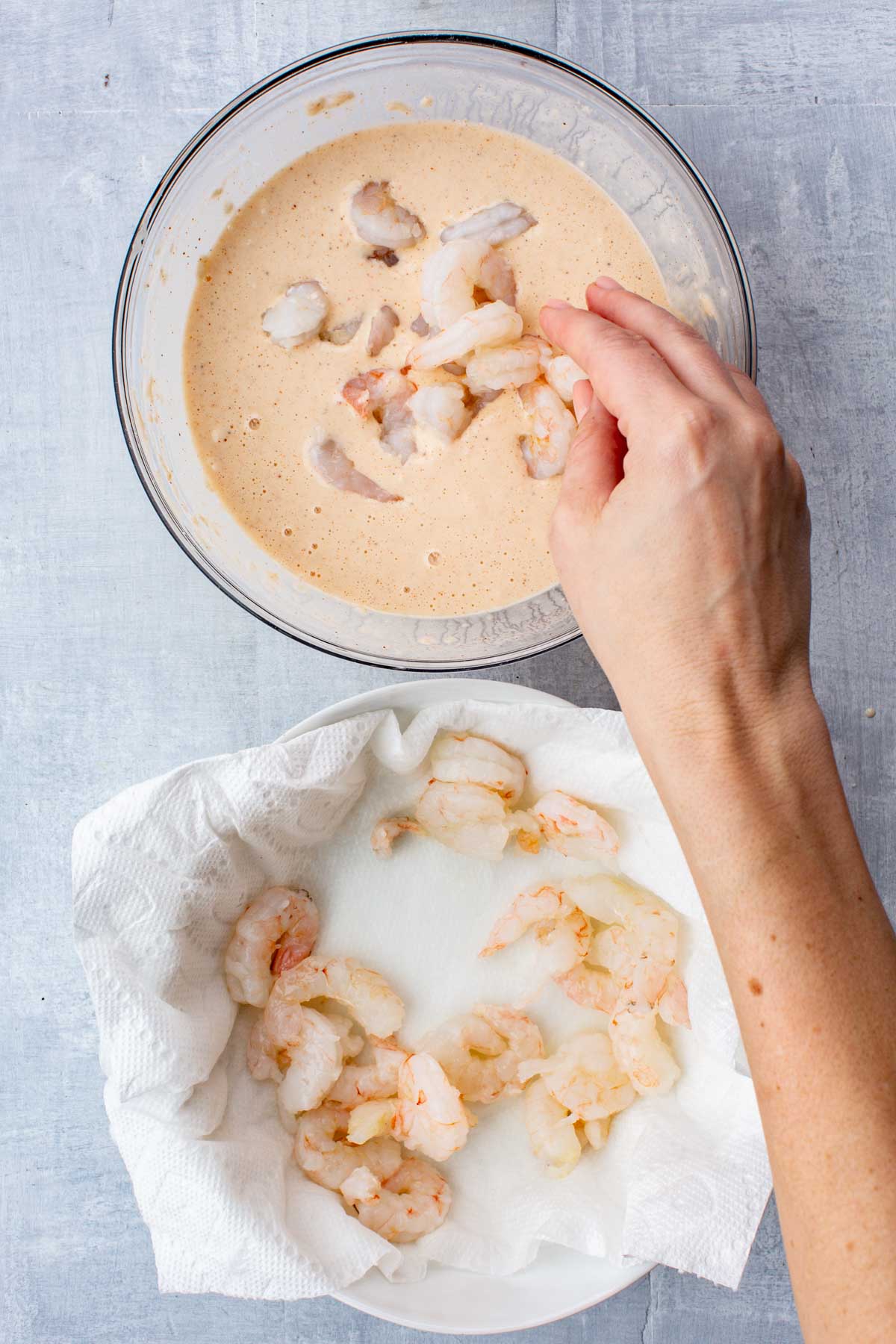 Hand placing raw shrimp into the beer batter, with more shrimp draining on a paper towel-lined plate.