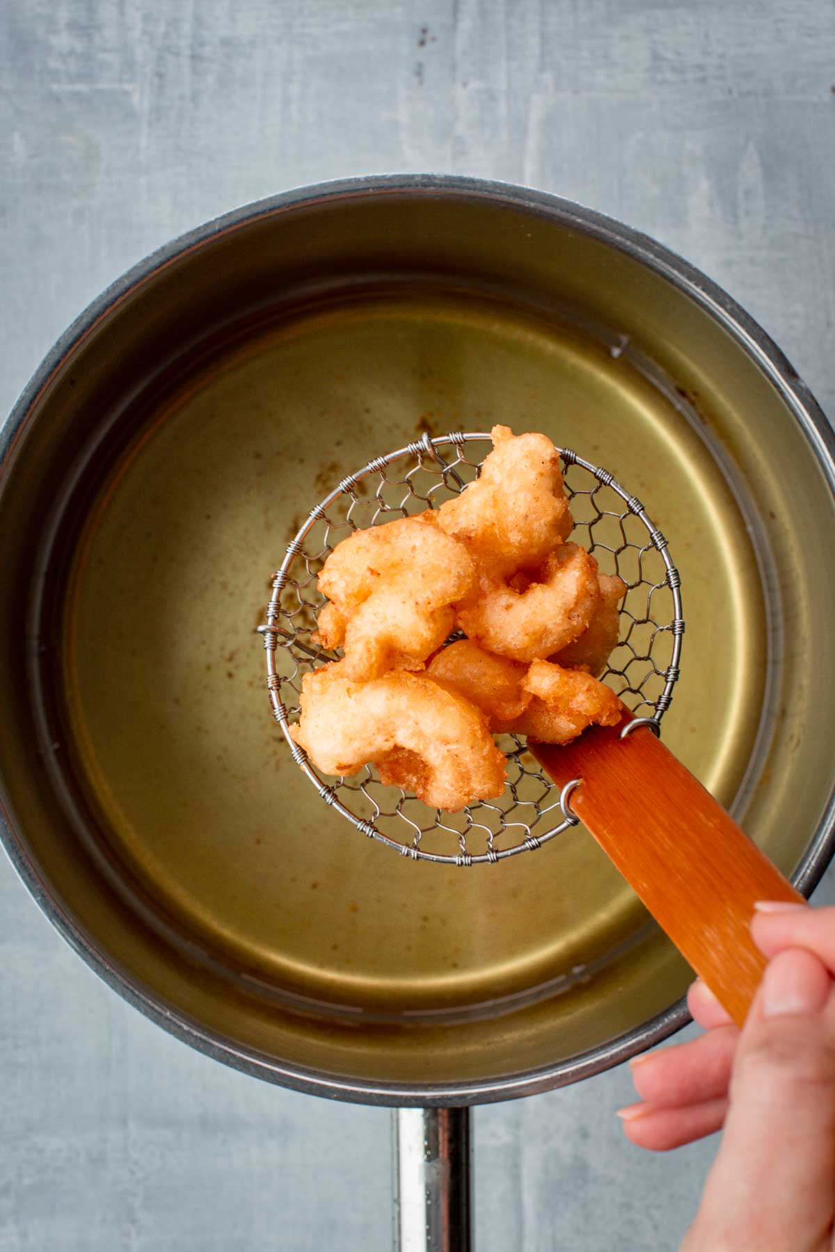 Fried shrimp being lifted from a pot of hot oil using a spider strainer with a wooden handle.