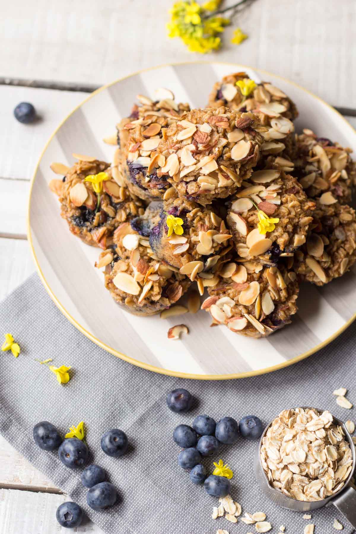 A striped plate full of oatmeal cups with blueberries and almonds, surrounded by oats, fresh blueberries, and yellow flowers.