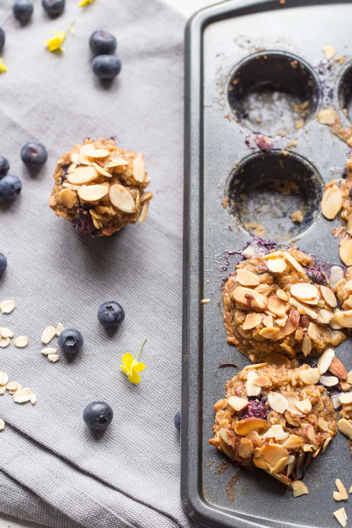 A single oatmeal cup sits next to a muffin tin with several missing and empty wells, surrounded by scattered oats and blueberries.