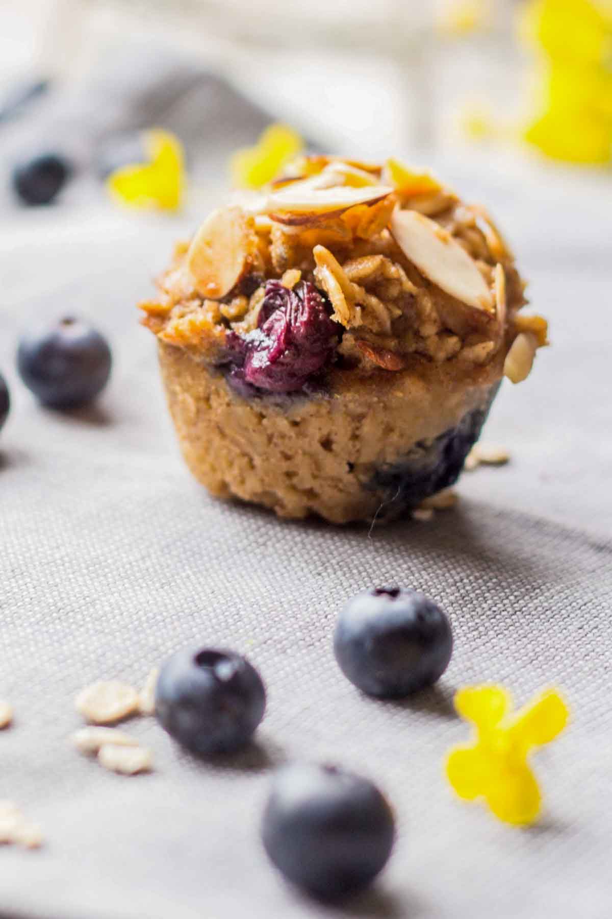 A close-up shot of a single oatmeal muffin topped with toasted almonds and juicy blueberries on a gray cloth background.