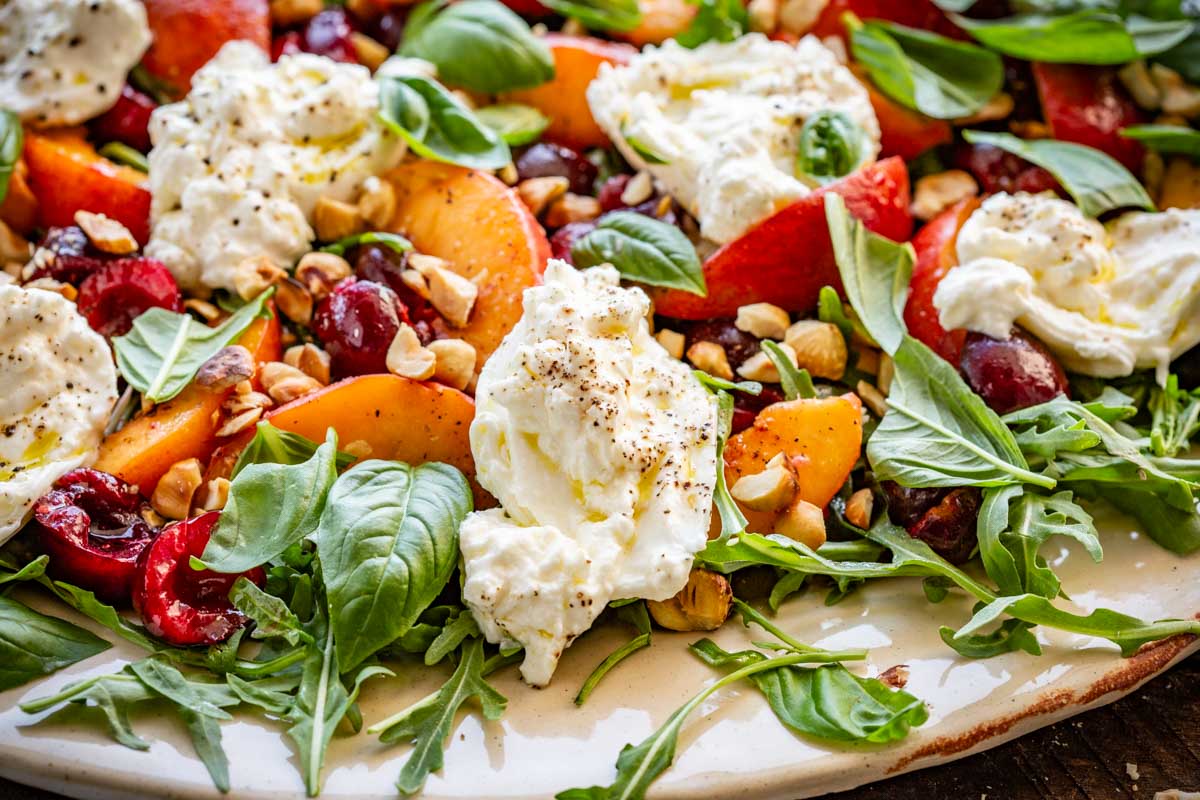 Detailed shot of a summer salad with creamy burrata, sliced peaches, cherries, toasted hazelnuts, fresh basil, and arugula on a rustic platter, seasoned with black pepper and drizzled with olive oil.