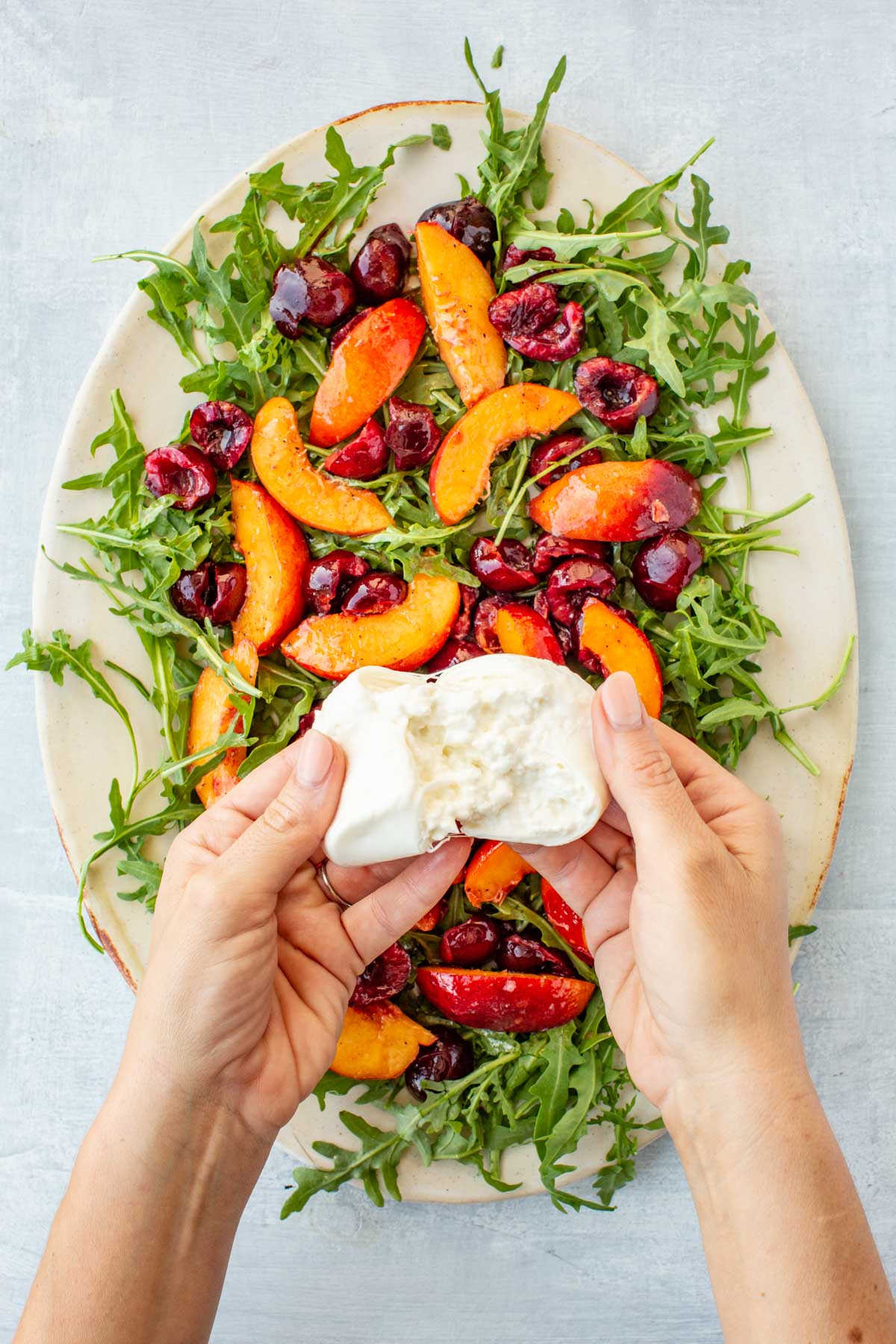 Sliced peaches and cherries scattered over the arugula base while hands hold burrata, ready to tear and add.