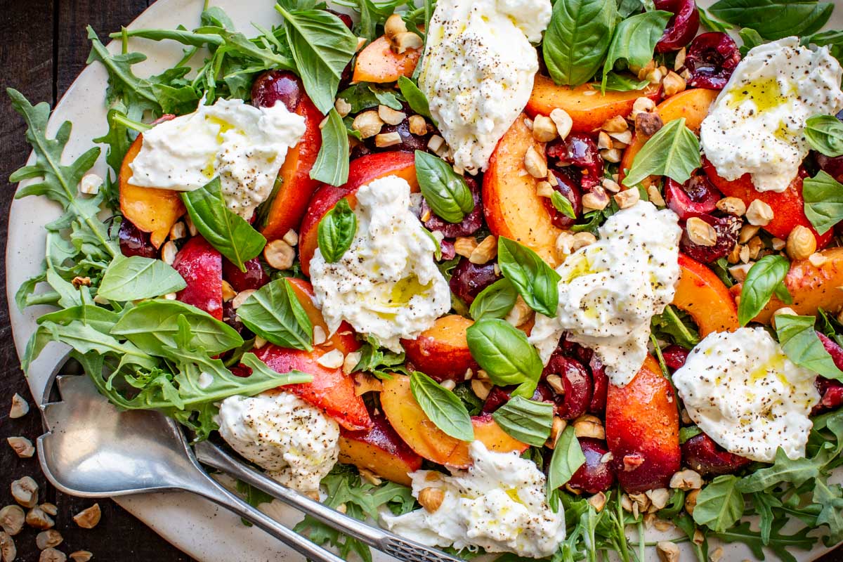 Close-up of the assembled salad with torn burrata, basil leaves, chopped hazelnuts, and fruit over arugula, drizzled with olive oil and cracked pepper.
