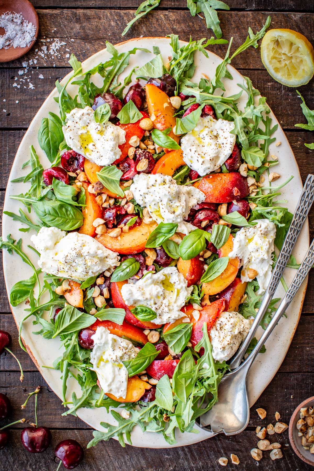 Full top-down view of the finished cherry peach burrata salad on a large platter with serving utensils and scattered ingredients.