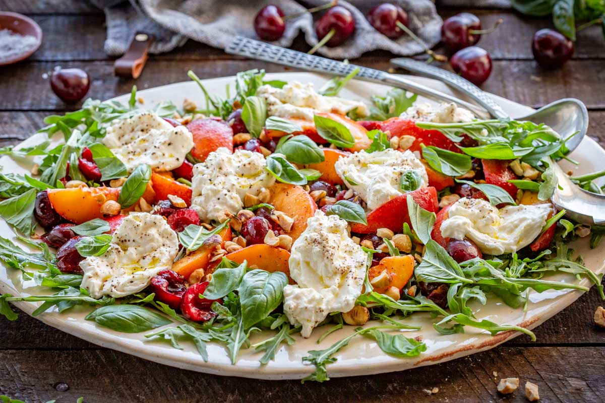 Side angle of the full salad platter showing volume and color with fresh basil, peaches, cherries, and burrata on a rustic wooden table.