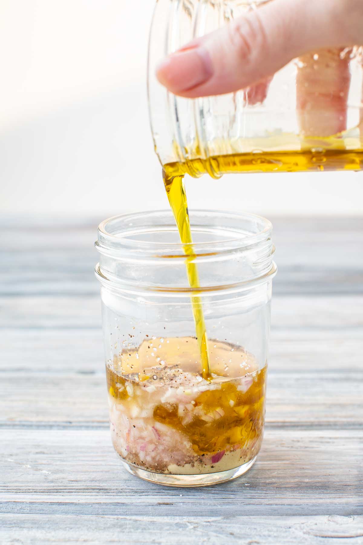 Olive oil being poured into a mason jar with minced shallots, mustard, vinegar, and seasonings to prepare a classic vinaigrette dressing.