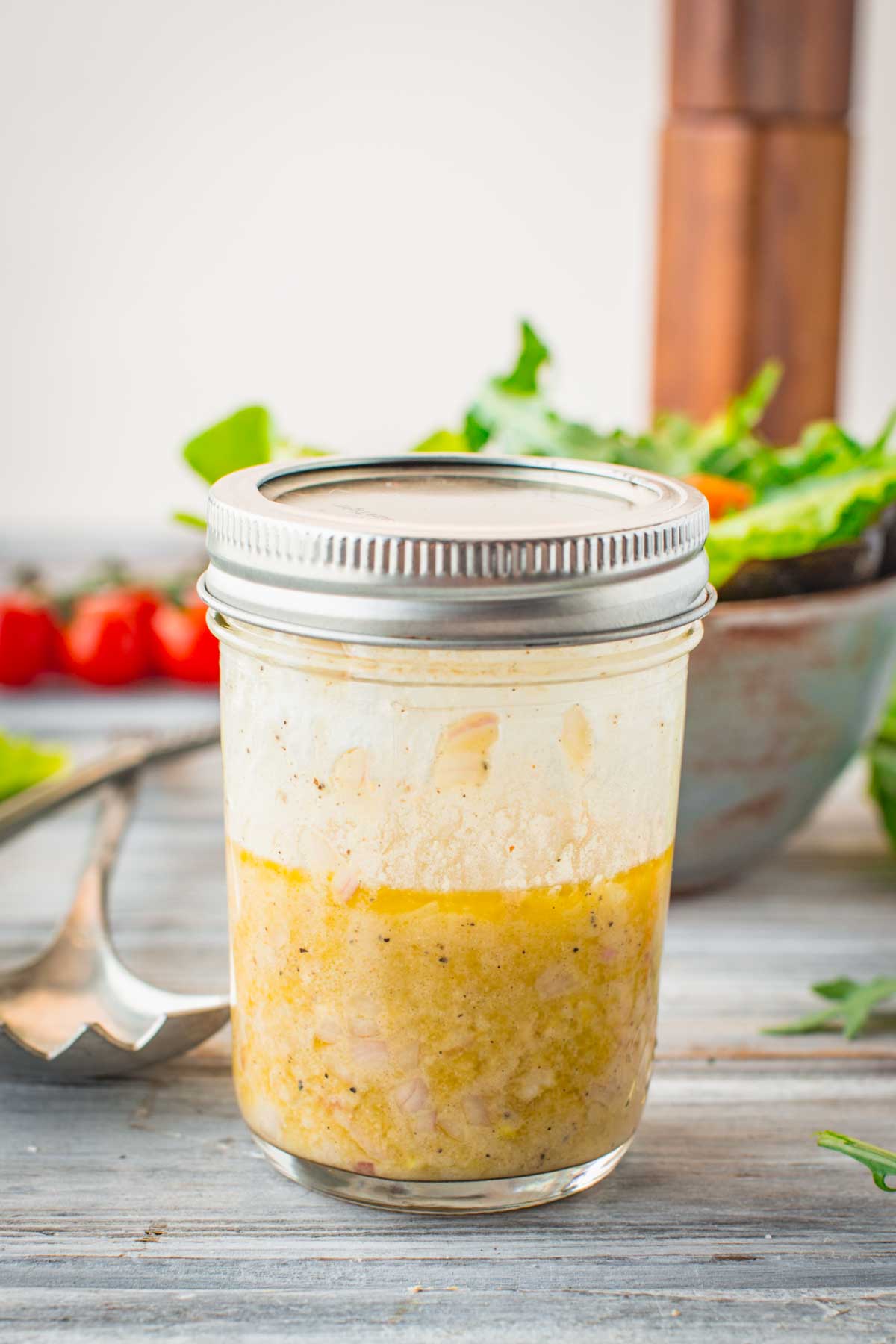 Homemade vinaigrette in a mason jar set on a rustic wooden surface with a salad and vegetables in the background, ready for serving.