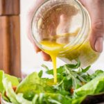 A hand pouring freshly made vinaigrette over a bowl of leafy green salad, with arugula and romaine in the foreground.