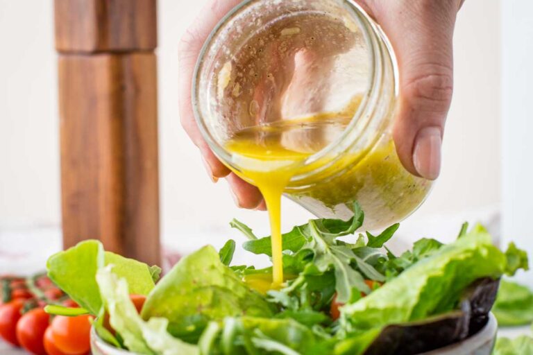 A hand pouring freshly made vinaigrette over a bowl of leafy green salad, with arugula and romaine in the foreground.