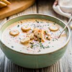 Creamy crab soup served in a green ceramic bowl, topped with oyster crackers, chopped herbs, and a sprinkle of paprika.