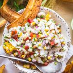 Top-down view of ceviche in a speckled bowl with spoon, surrounded by fresh cilantro, corn on the cob, and long fried plantain chips.