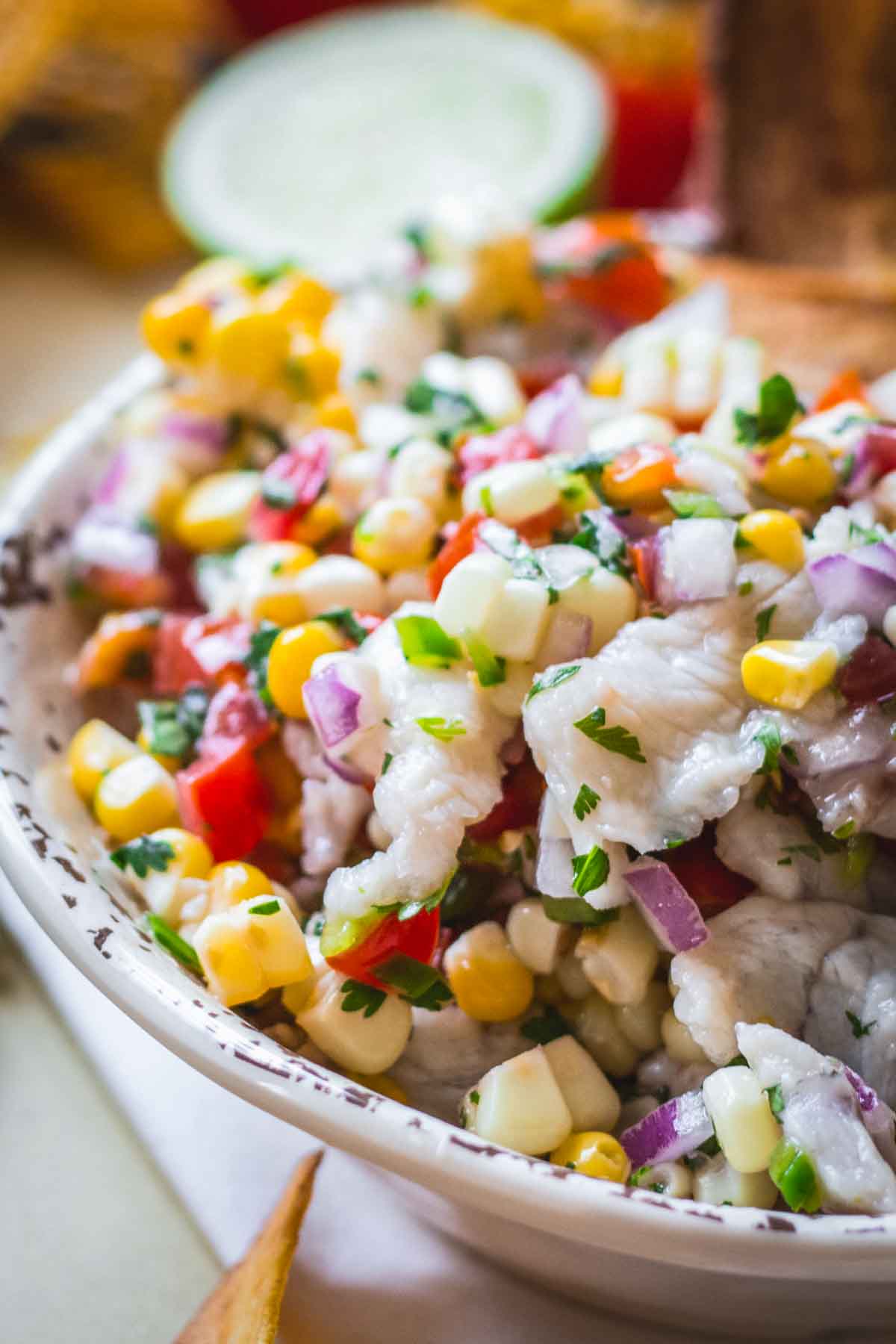 Bright and fresh fish ceviche with sweet corn, red onion, tomatoes, and cilantro in a rustic ceramic bowl with lime in the background.
