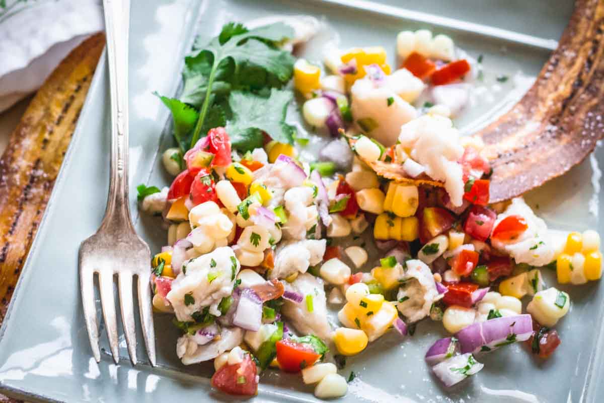 Fork resting on a plate of partially eaten fish ceviche with corn, red onion, tomato, and herbs, served with fried plantain chips.