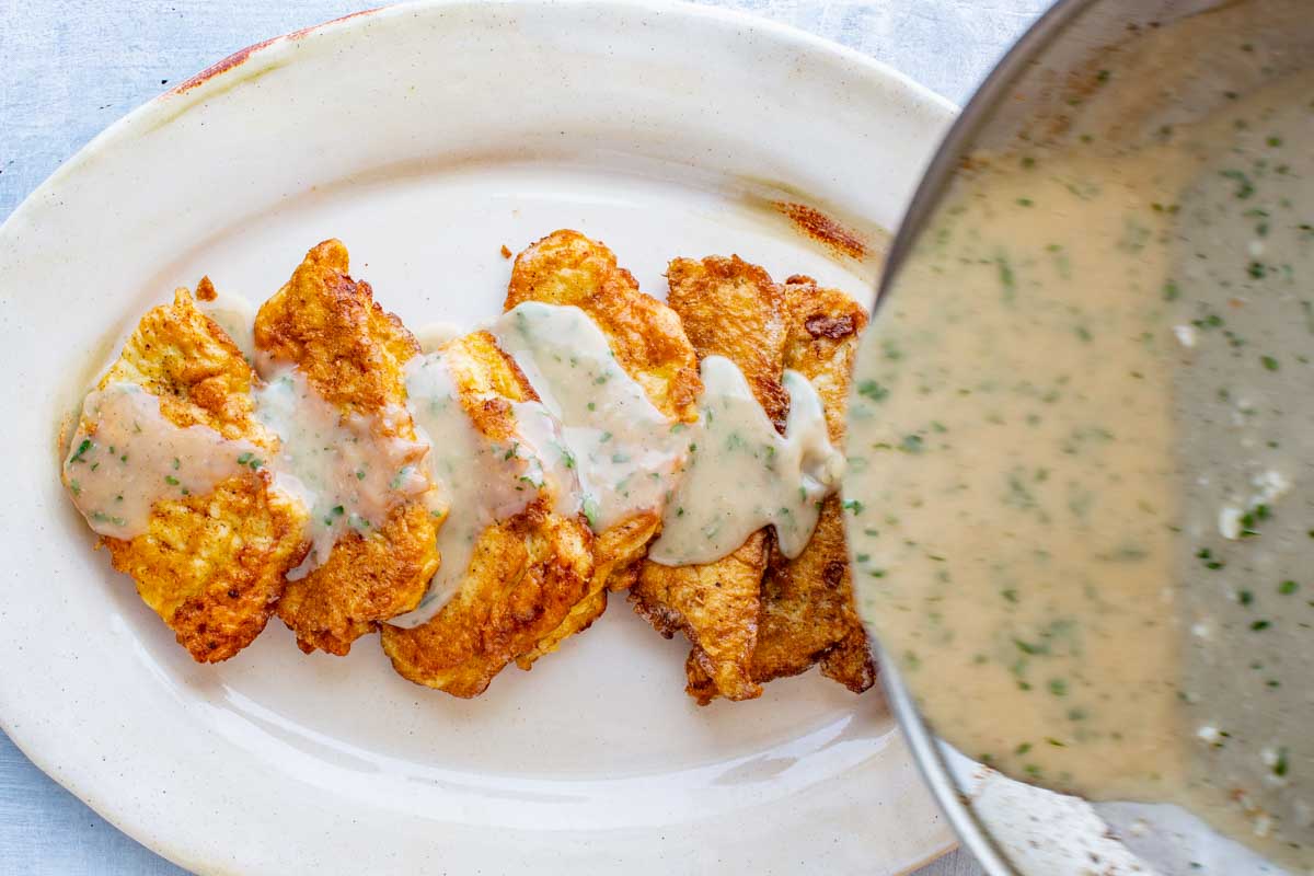 Golden-brown flounder filets lined up on a white platter with creamy lemon herb sauce being poured over top.