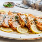 Close-up of crispy flounder francaise covered in silky lemon sauce, garnished with chopped parsley and lemon slices.