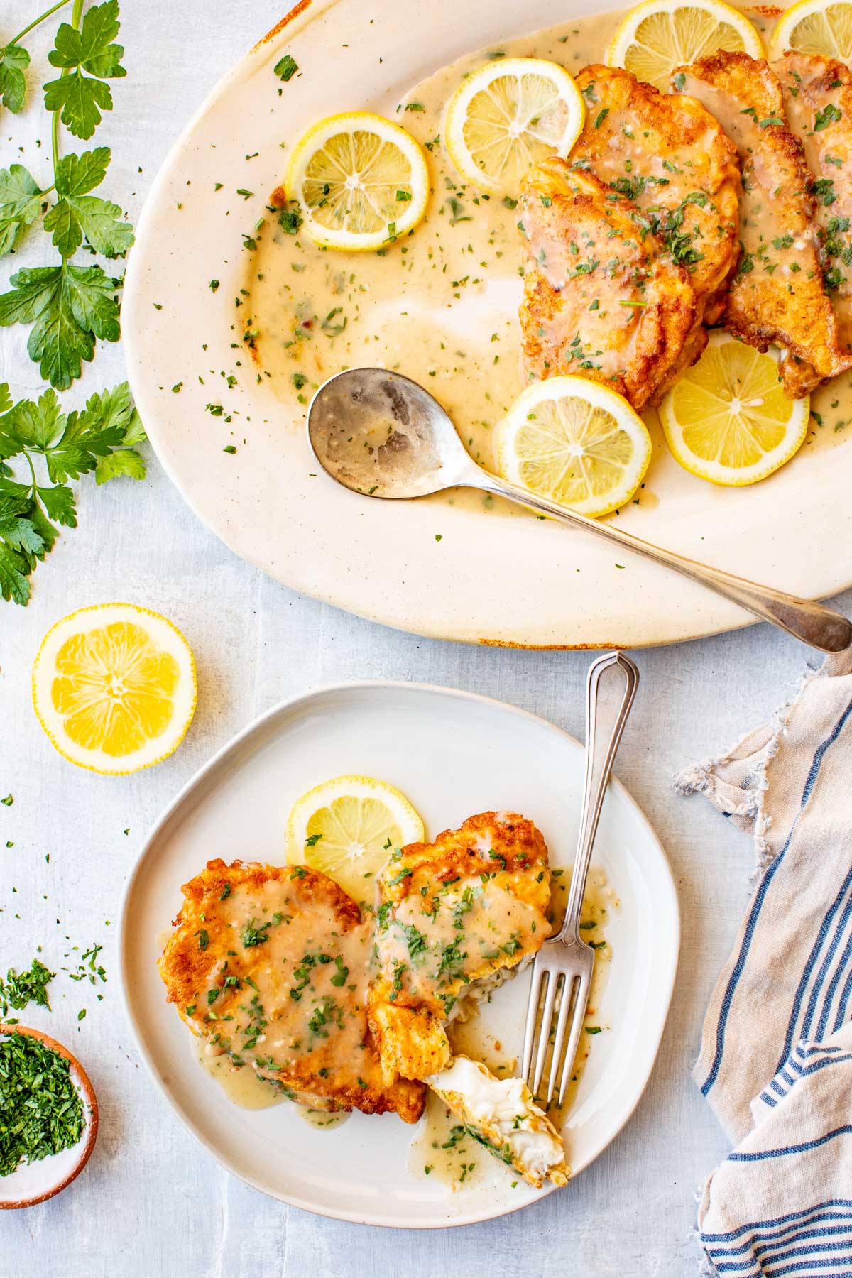 Partial serving platter of flounder francaise next to a small plate with two pieces served alongside lemon slices and a fork.