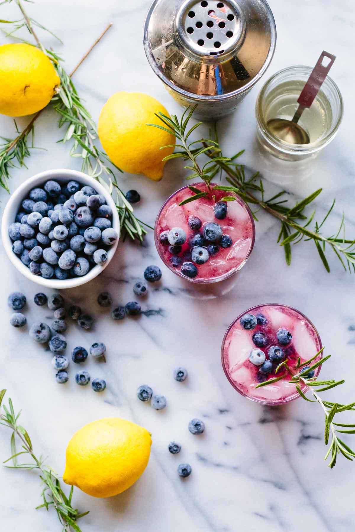 Overhead shot of blueberries, lemons, rosemary, a cocktail shaker, simple syrup, and two blueberry rosemary vodka spritzers garnished with fresh berries and rosemary.