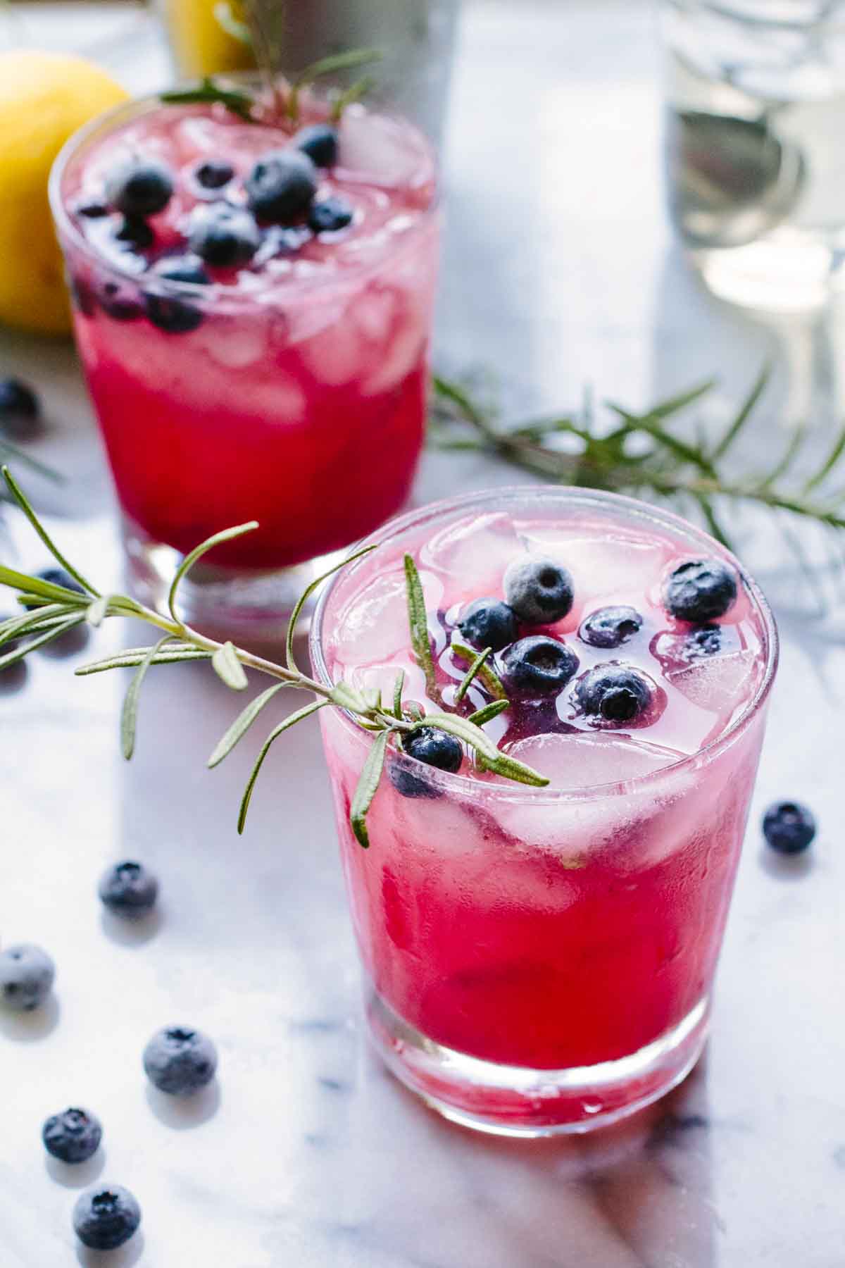 Close-up of two glasses filled with pink blueberry rosemary spritzers, ice, whole blueberries, and a sprig of rosemary for garnish.
