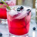 Glass of blueberry rosemary vodka spritzer on a marble surface, topped with ice, blueberries, and a rosemary sprig, with another glass in the background.