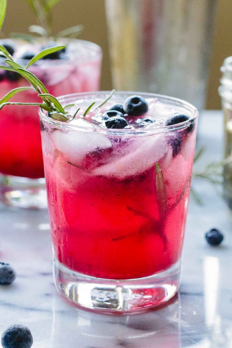 Glass of blueberry rosemary vodka spritzer on a marble surface, topped with ice, blueberries, and a rosemary sprig, with another glass in the background.