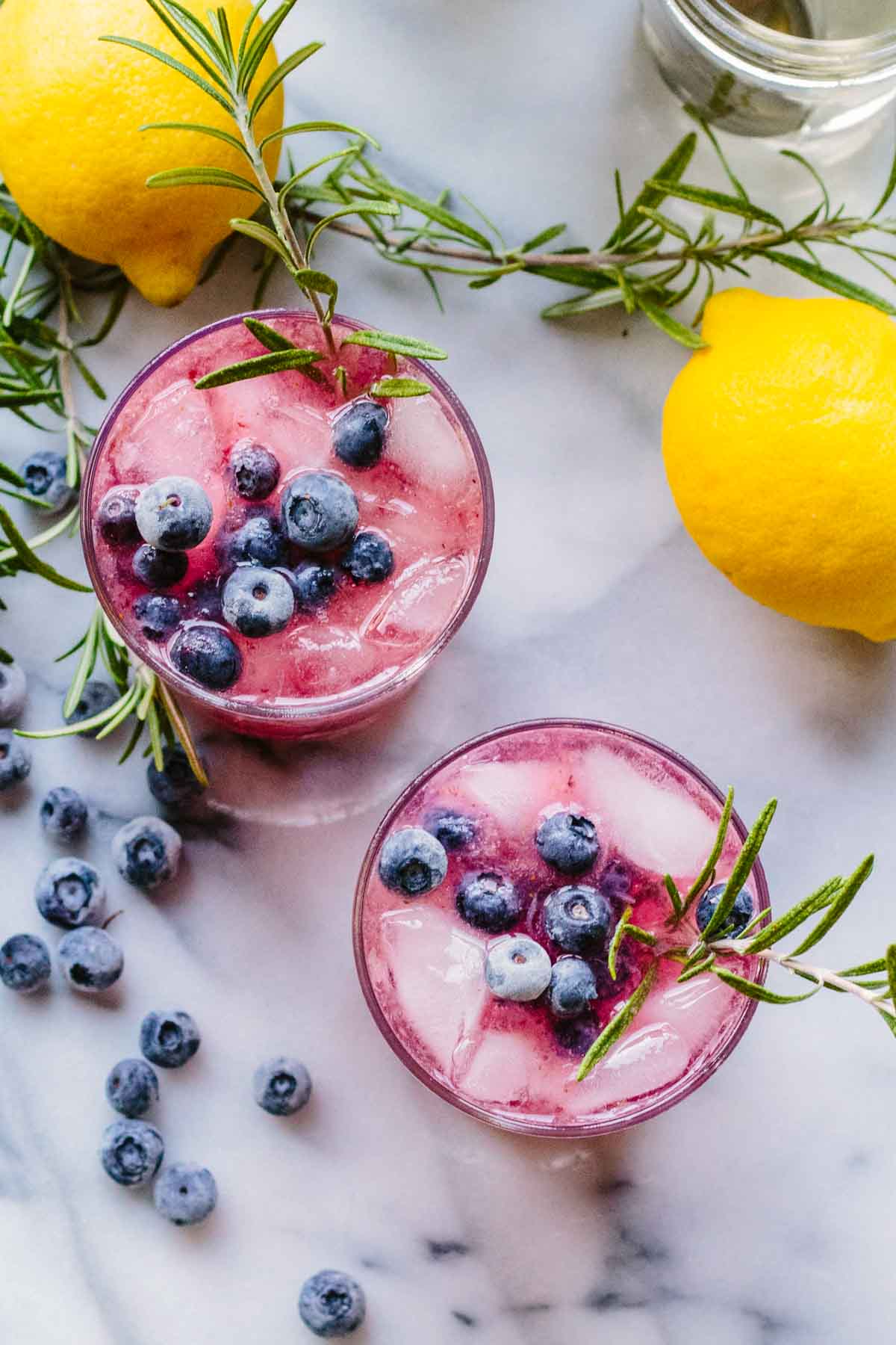 Overhead view of two spritzers garnished with rosemary, surrounded by loose blueberries, whole lemons, and rosemary sprigs on a marble background.