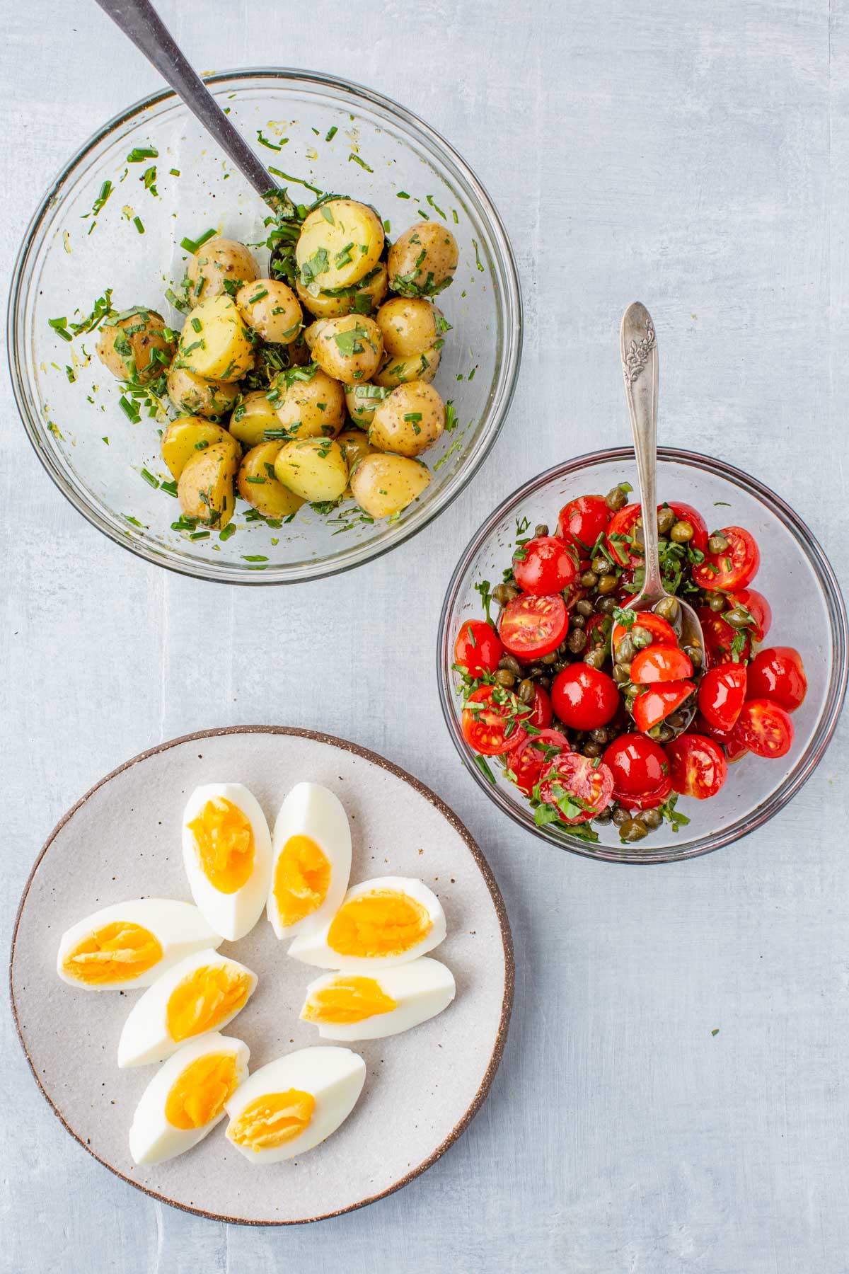 Three separate bowls containing halved herbed potatoes, cherry tomato and caper salad, and halved soft-boiled eggs on a white background.