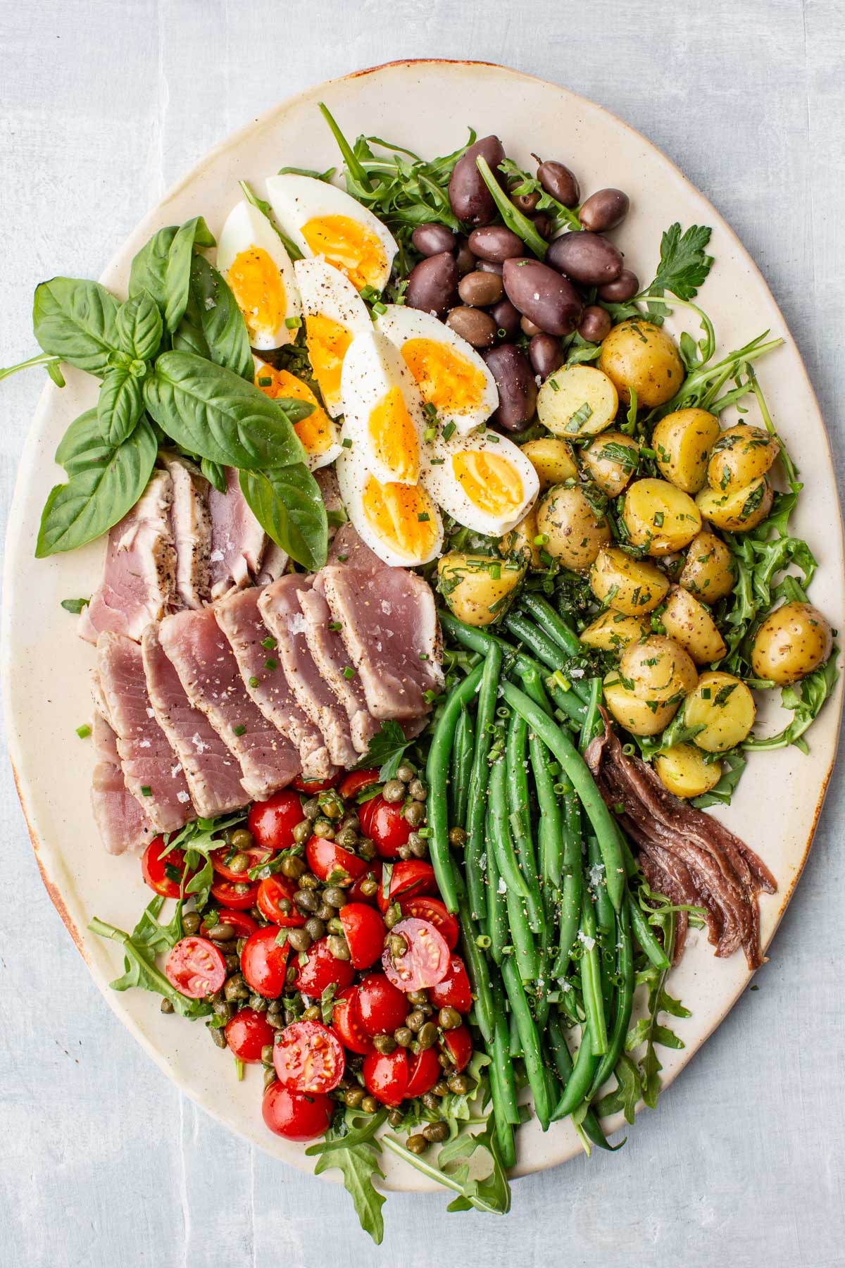 Beautifully arranged Niçoise salad on a white platter with sliced seared tuna, green beans, olives, herbed potatoes, cherry tomatoes with capers, soft-boiled eggs, anchovies, and fresh basil.