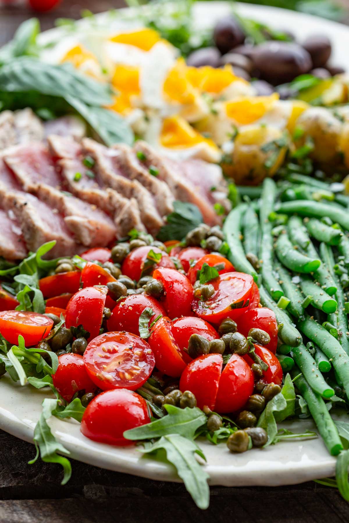 Close-up of glossy cherry tomatoes and briny capers over arugula and next to green beans and sliced tuna.