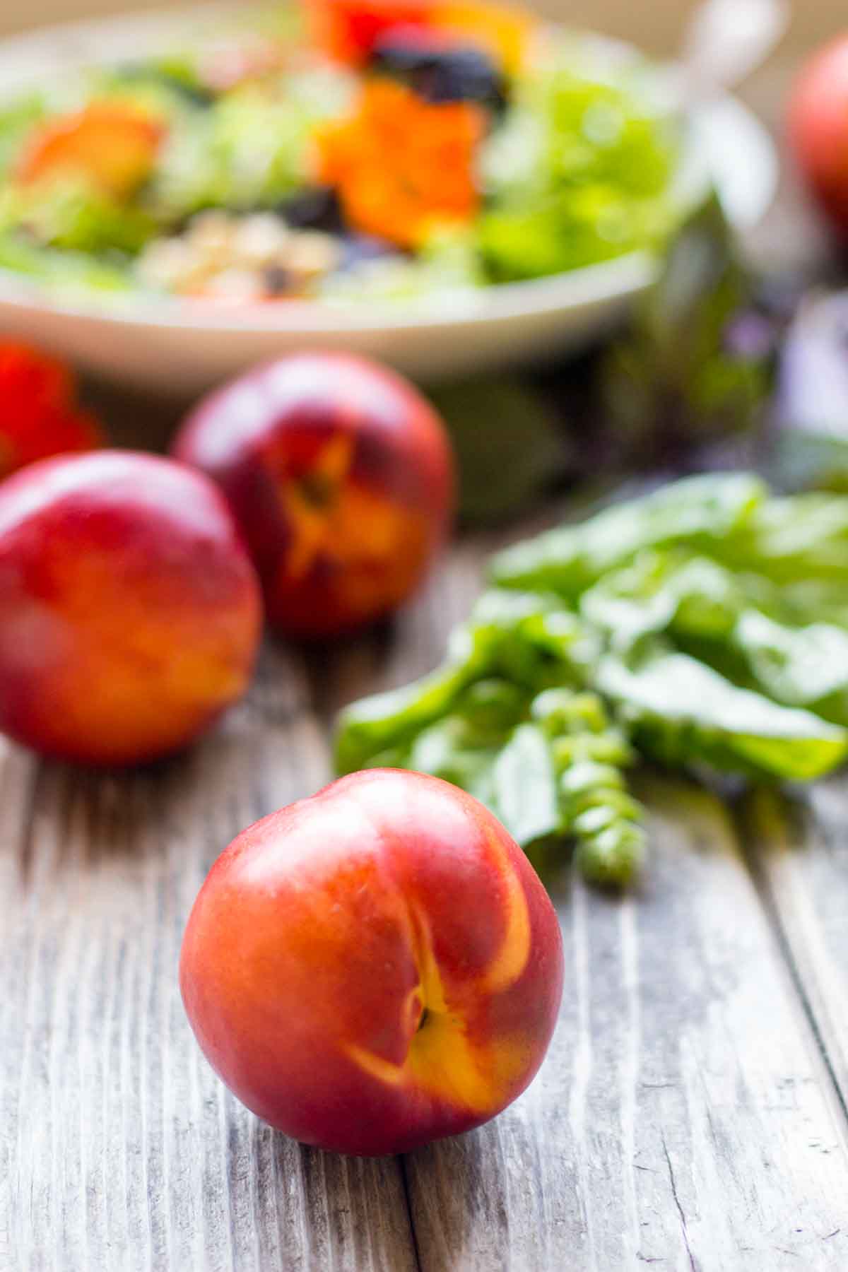 Whole nectarines and fresh basil leaves on a rustic wooden surface with a blurred summer salad in the background.