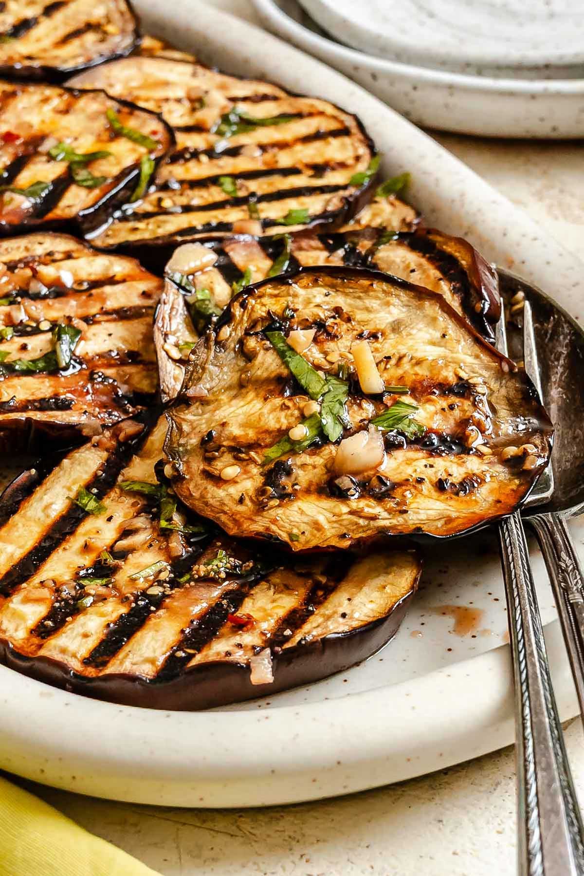 Side angle of the serving platter filled with marinated grilled eggplant, next to a stack of ceramic plates.